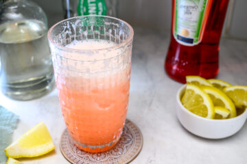 A glass of pink cocktail with frothy top sits on a coaster, surrounded by bottles and a bowl of lemon wedges on a marble countertop.