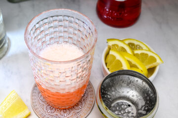 A textured glass with a light pink frothy drink sits on a coaster beside a metal strainer and a small bowl of lemon slices on a marble surface.