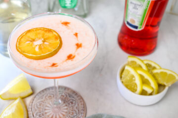 A cocktail in a coupe glass garnished with a dried orange slice, surrounded by lemon wedges, a bottle of Aperitivo, and a green bottle on a white surface.