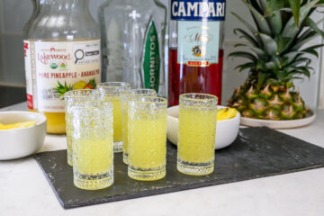 Four filled shot glasses of yellow liquid are placed on a slate tray, with bottles of pineapple juice, tequila, Campari, a pineapple, and a small bowl of pineapple slices nearby.