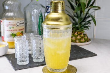 A gold cocktail shaker filled with a yellow drink sits on a coaster, with empty patterned glasses and bottles of juice and liquor in the background.