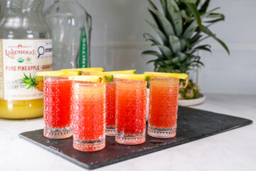 Five red and orange layered cocktails in patterned glasses, each garnished with a pineapple slice, are arranged on a black tray with a pineapple and juice bottles in the background.