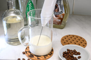 A hand blender is mixing milk in a glass measuring jug on a kitchen counter, surrounded by bottles of alcohol, a coaster, and a dish with coffee beans.
