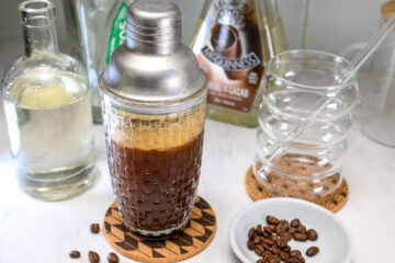 A cocktail shaker filled with a dark beverage sits on a coaster, surrounded by bottles, empty glasses, a glass straw, and coffee beans on a small plate.
