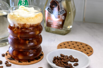 A glass of iced coffee topped with whipped cream sits on a coaster next to a bowl of coffee beans and liquor bottles in the background.