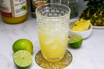 A glass of yellow beverage with ice sits on a coaster, surrounded by a halved lime, a whole lime, pineapple slices, and bottles in the background.