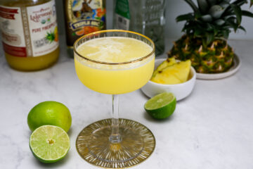 A cocktail glass filled with a yellow tropical drink sits on a counter with limes, pineapple slices, a whole pineapple, and bottles of juice and alcohol in the background.