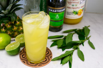 A tall glass of yellow-green drink with ice sits on a coaster, surrounded by fresh lime, mint leaves, bottles of Chartreuse, pineapple juice, and a whole pineapple.
