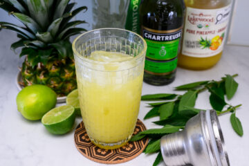 A glass of yellow cocktail with ice sits on a coaster, surrounded by a pineapple, limes, a cocktail shaker, mint leaves, and bottles of Chartreuse and pineapple juice.