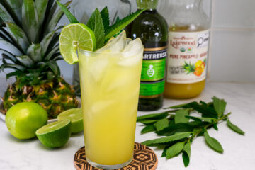 A glass of yellow cocktail with ice, lime slices, and mint leaves, placed in front of a pineapple, limes, pineapple juice, and a green liqueur bottle on a white counter.