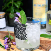 A cocktail in a glass with black salt on the rim, filled with ice and garnished with a sage leaf and purple flowers, with bottles and plants in the background.