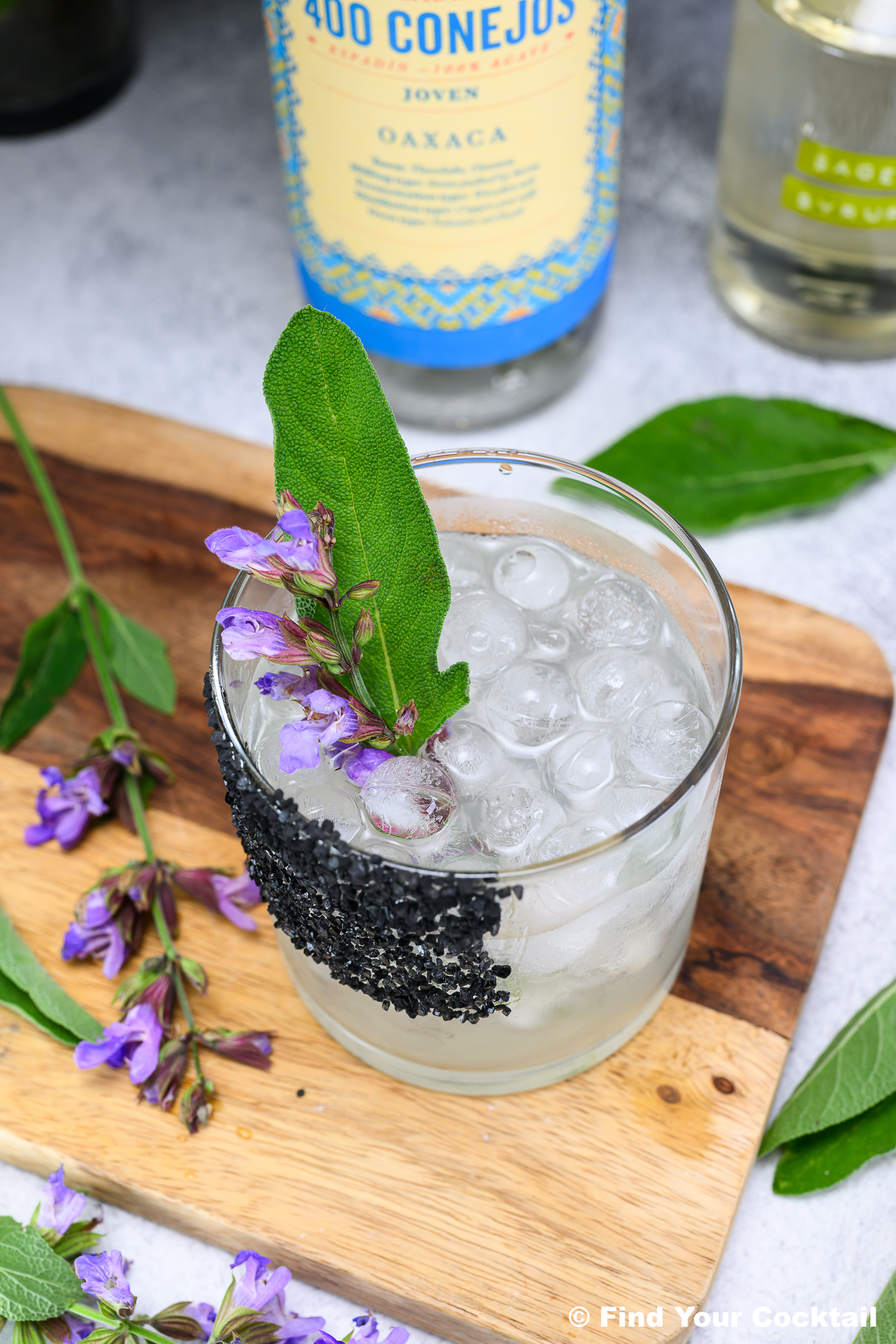A cocktail with ice in a glass, garnished with a sage leaf and purple flowers, with a black salt rim, sits on a wooden board. A bottle labeled 400 Conejos is in the background.