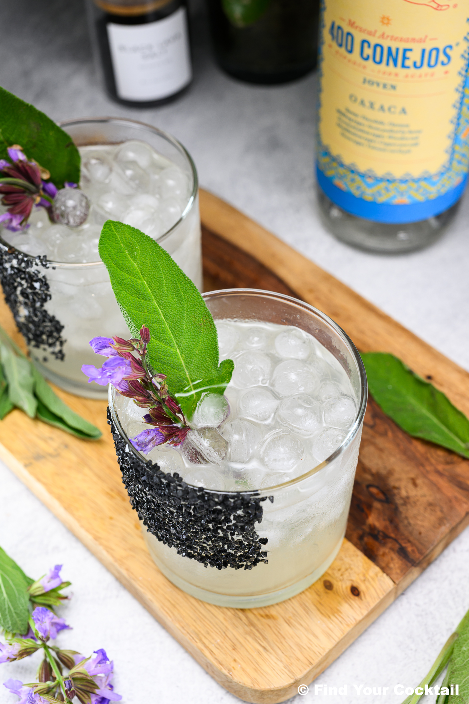 Two cocktails with ice, black salt on the rim, and garnished with a green leaf and purple flowers are placed on a wooden tray. A bottle labeled 400 Conejos is nearby.