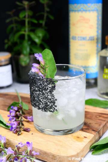A cocktail in a glass with black salt on the rim, filled with ice and garnished with a sage leaf and purple flowers, with bottles and plants in the background.