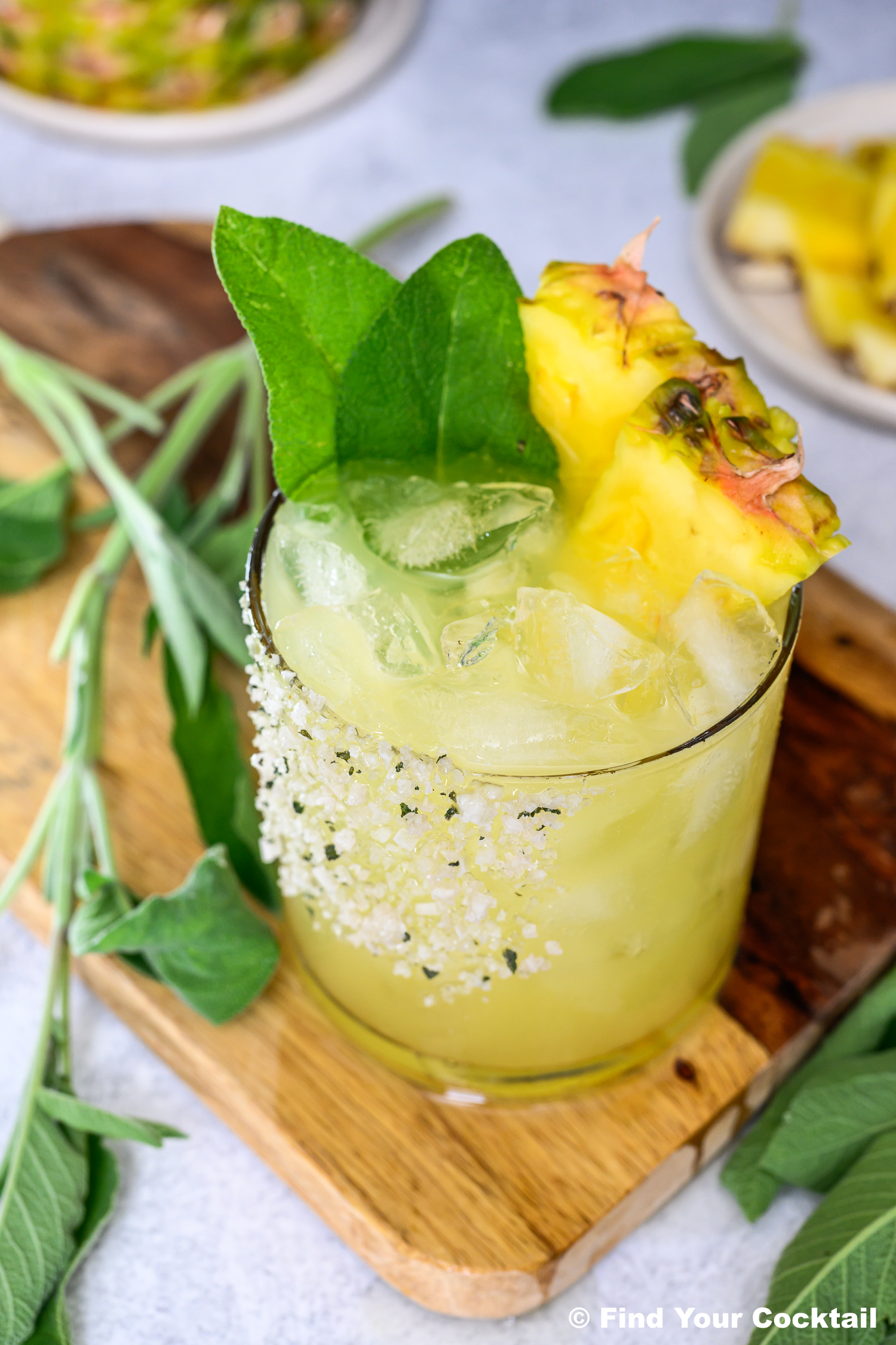 A glass of yellow cocktail with ice, rimmed with salt, garnished with pineapple slices and fresh green leaves, placed on a wooden board with herbs nearby.