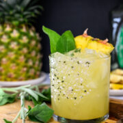 A glass of yellow cocktail with ice, garnished with pineapple slices and green sage leaves, sits on a wooden board near fresh sage and a whole pineapple.