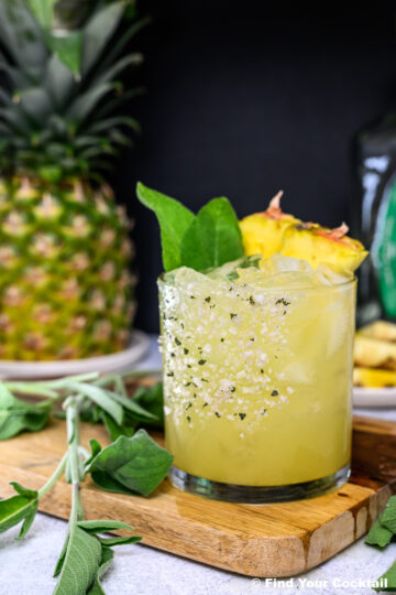 A glass of yellow cocktail with ice, garnished with pineapple slices and green sage leaves, sits on a wooden board near fresh sage and a whole pineapple.