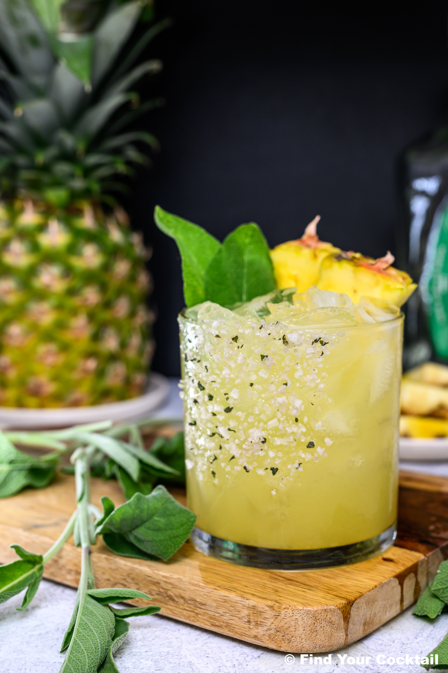 A glass of yellow cocktail with ice, garnished with pineapple slices and green sage leaves, sits on a wooden board near fresh sage and a whole pineapple.