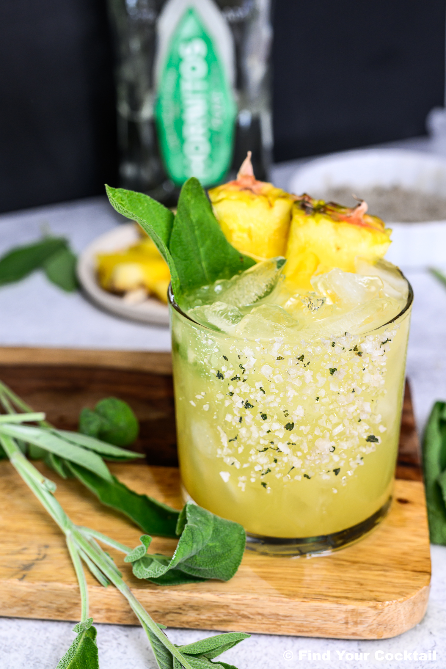 A glass of yellow cocktail with ice, pineapple slices, green herbs, and a salt rim sits on a wooden board, with fresh sage and a bottle in the background.