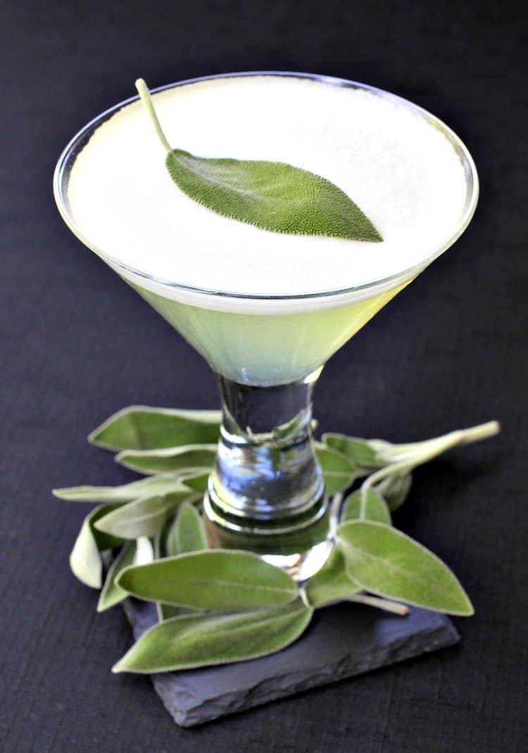 A cocktail in a martini glass topped with a single sage leaf, garnished with additional sage leaves on a slate coaster.