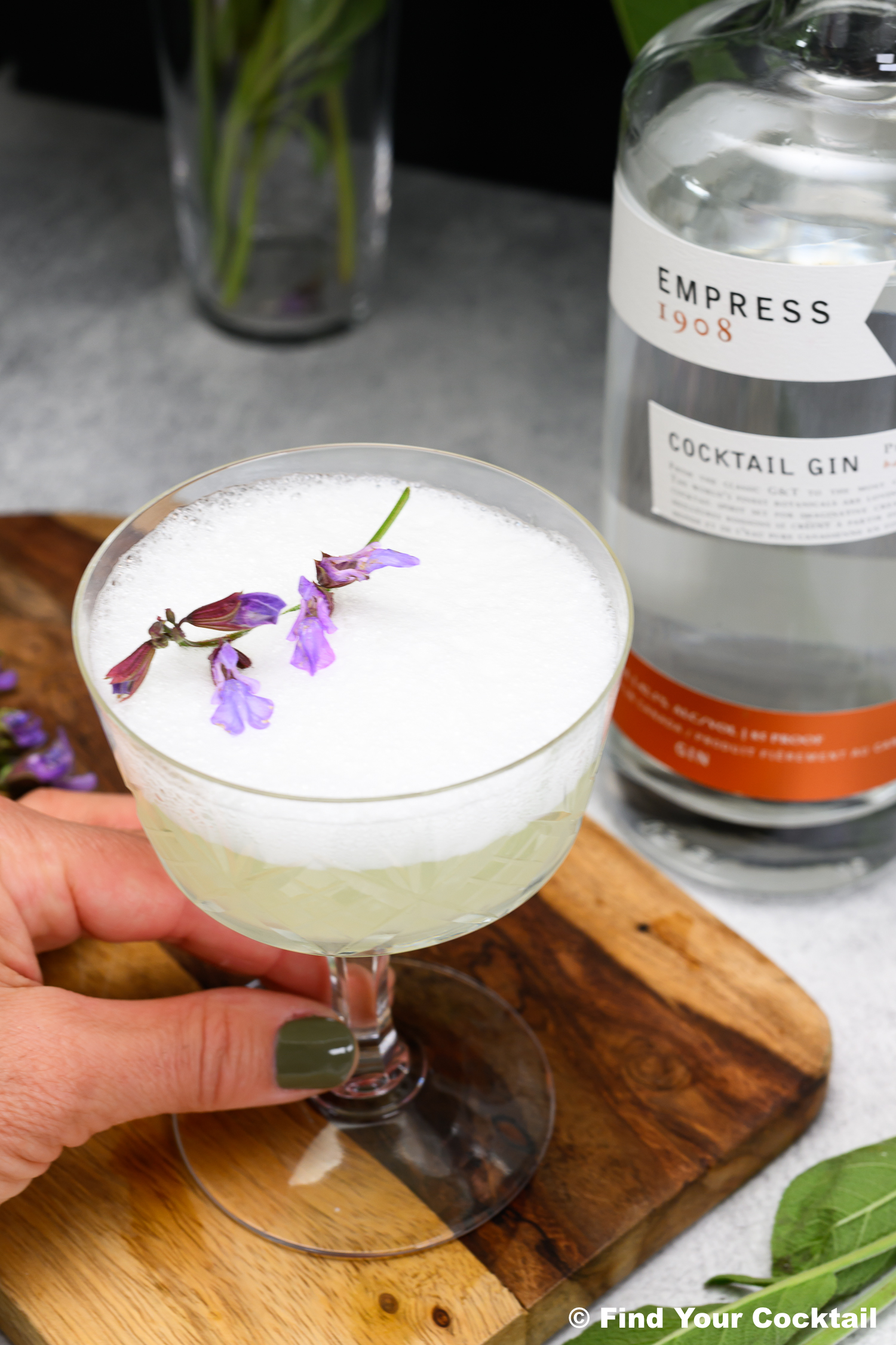 A hand holds a coupe glass with a frothy cocktail garnished with purple flowers, next to a bottle of Empress 1908 gin on a wooden board.