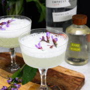 Two coupe glasses filled with a frothy, pale cocktail garnished with purple flowers, placed on a wooden board with gin and sage liqueur bottles in the background.