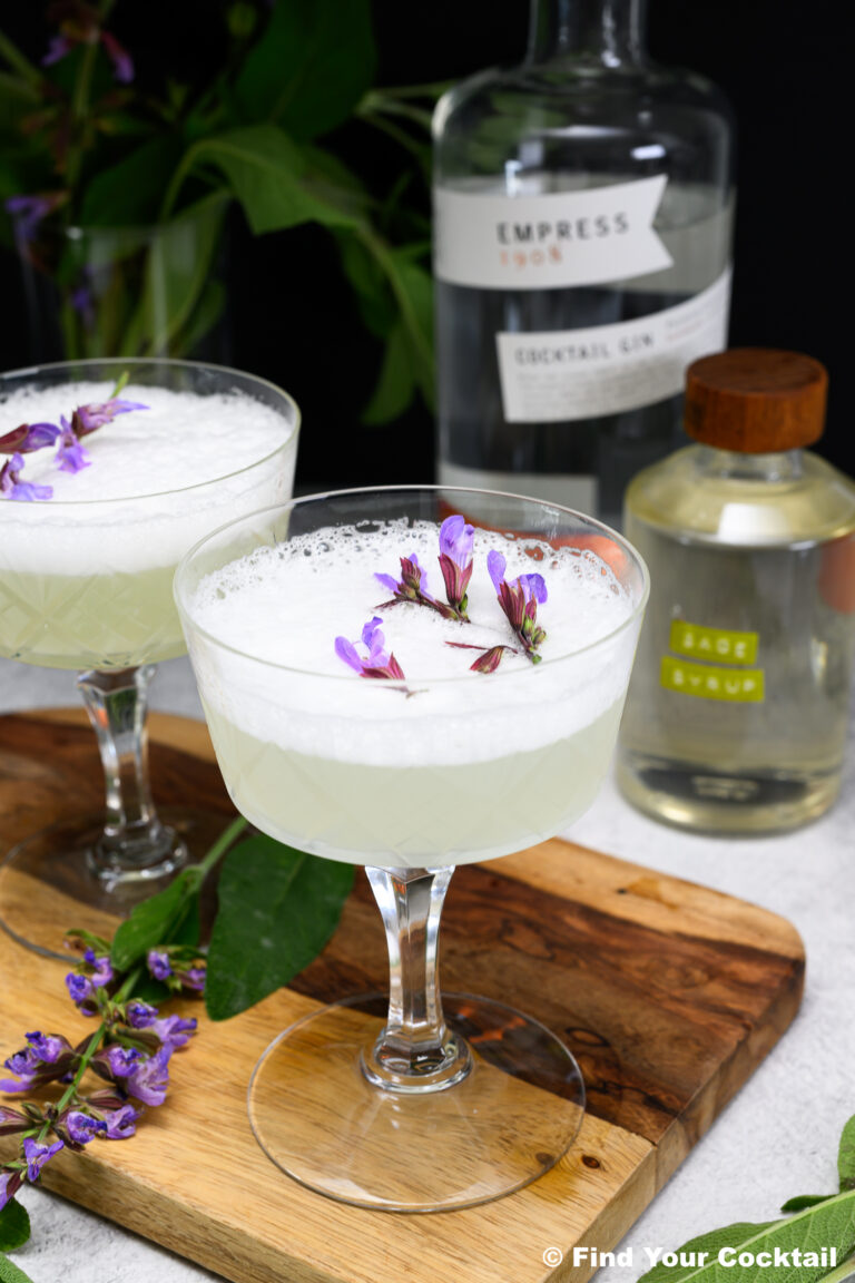 Two coupe glasses filled with a frothy, pale cocktail garnished with purple flowers, placed on a wooden board with gin and sage liqueur bottles in the background.