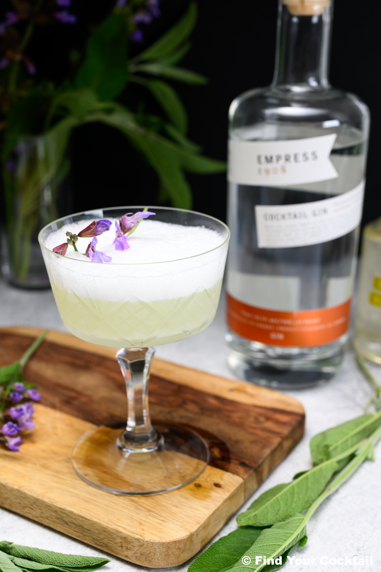 A gin cocktail with a frothy top, garnished with purple flowers, sits on a wooden board beside a bottle of Empress 1908 gin and green leaves.