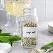 A glass jar labeled sage salt sits on a counter with a spoonful of the salt, fresh sage leaves, and two crystal cocktail glasses in the background.