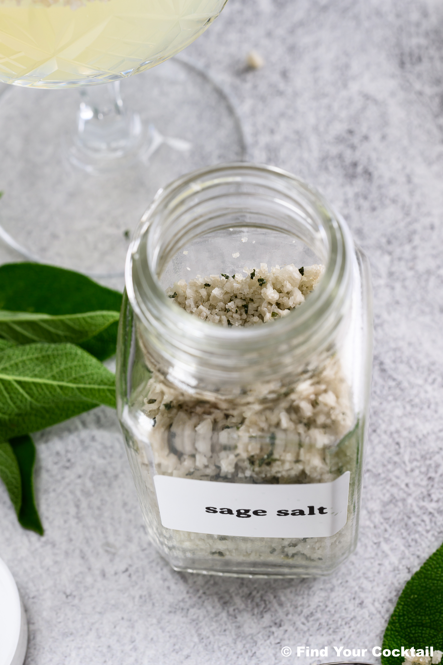 A glass jar labeled sage salt filled with coarse salt and sage sits on a gray surface next to fresh sage leaves.