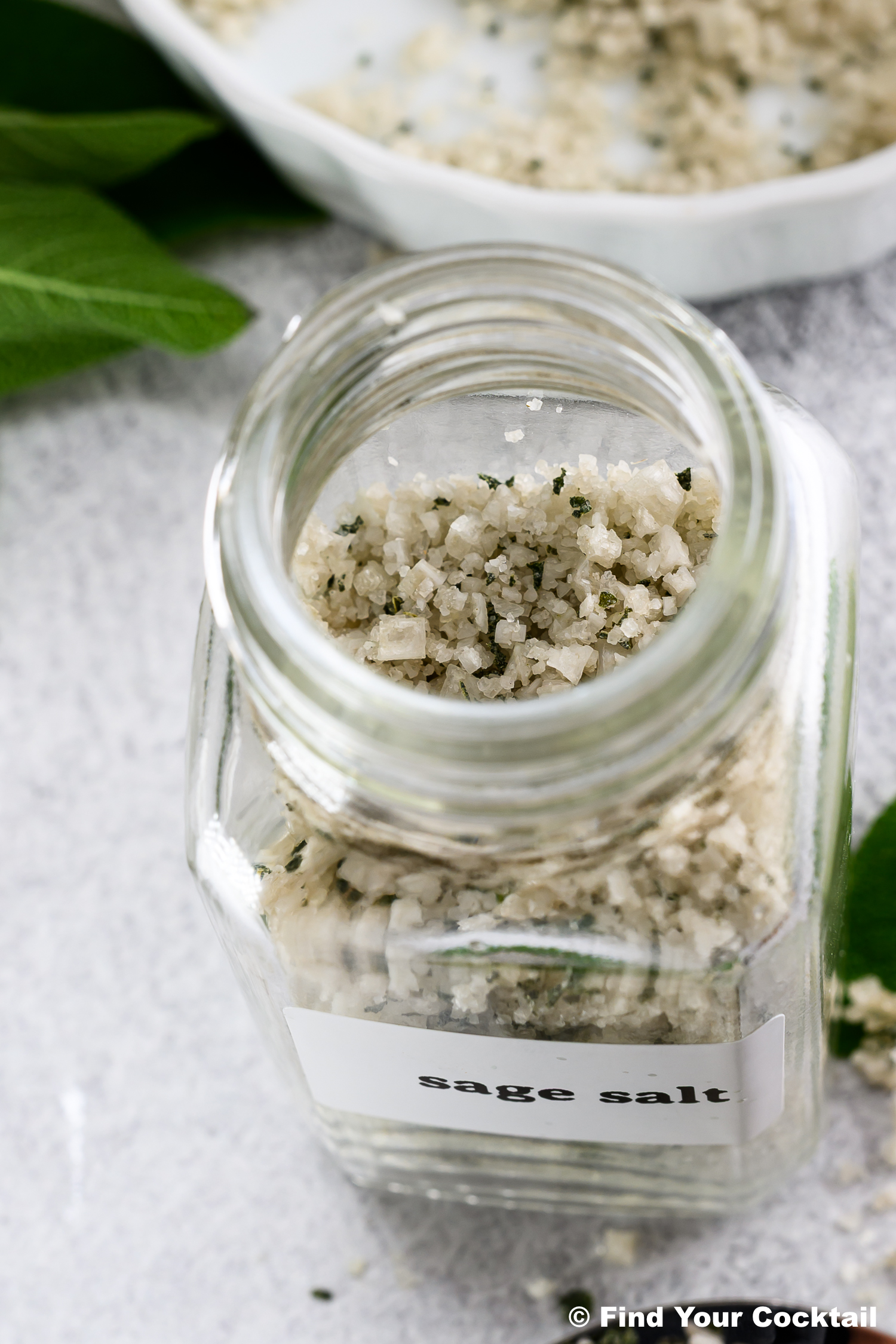 A glass jar labeled sage salt filled with coarse salt mixed with dried sage leaves, with some sage salt and fresh sage leaves in the background.