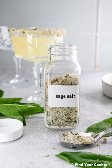 A glass jar labeled sage salt sits on a counter with a spoonful of the salt, fresh sage leaves, and two crystal cocktail glasses in the background.