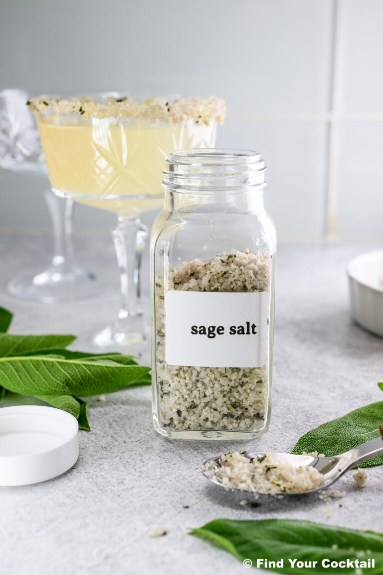 A glass jar labeled sage salt sits on a counter with a spoonful of the salt, fresh sage leaves, and two crystal cocktail glasses in the background.