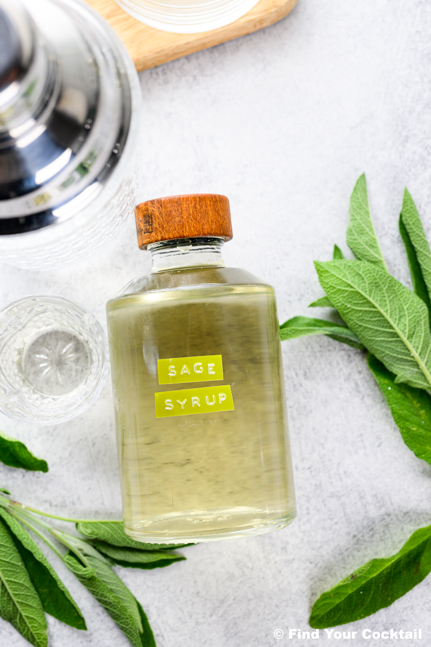 A glass bottle labeled Sage Syrup with a wooden cap sits next to a cocktail shaker, empty glass, and fresh sage leaves on a light surface.
