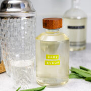 A glass bottle labeled Sage Simple Syrup with a wooden cap is on a counter next to a glass, a cocktail shaker, another bottle, and fresh sage leaves.