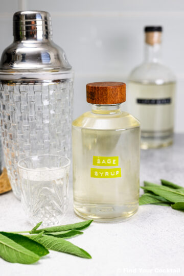 A glass bottle labeled Sage Simple Syrup with a wooden cap is on a counter next to a glass, a cocktail shaker, another bottle, and fresh sage leaves.