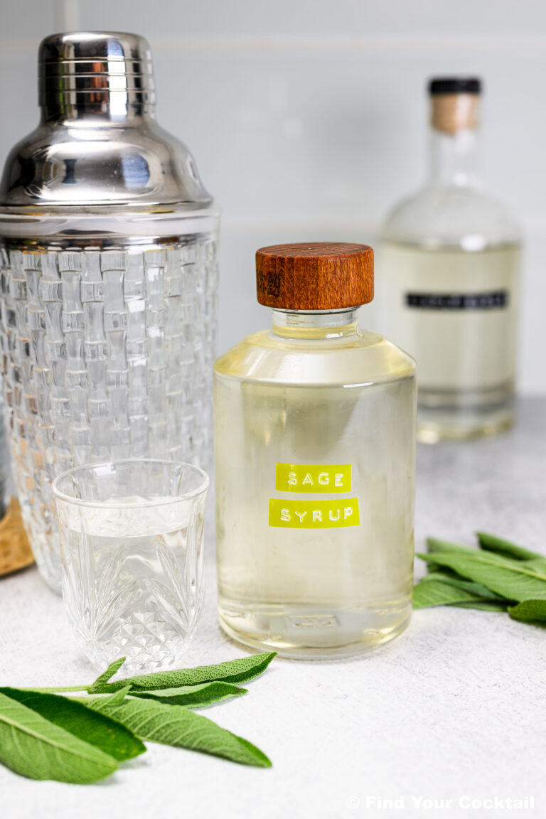 A glass bottle labeled Sage Simple Syrup with a wooden cap is on a counter next to a glass, a cocktail shaker, another bottle, and fresh sage leaves.