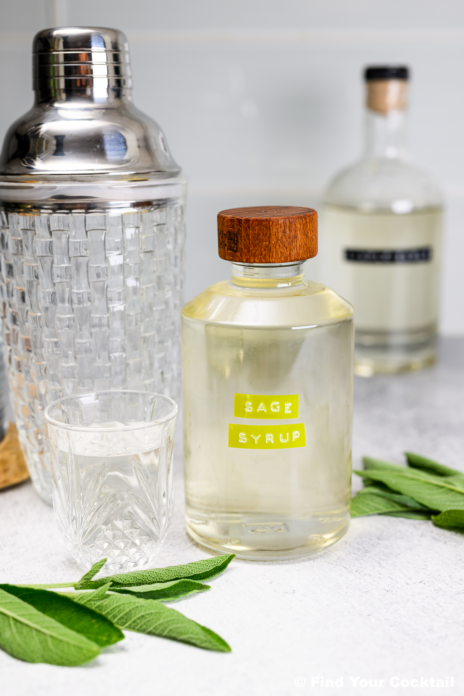 A glass bottle labeled Sage Simple Syrup with a wooden cap is on a counter next to a glass, a cocktail shaker, another bottle, and fresh sage leaves.