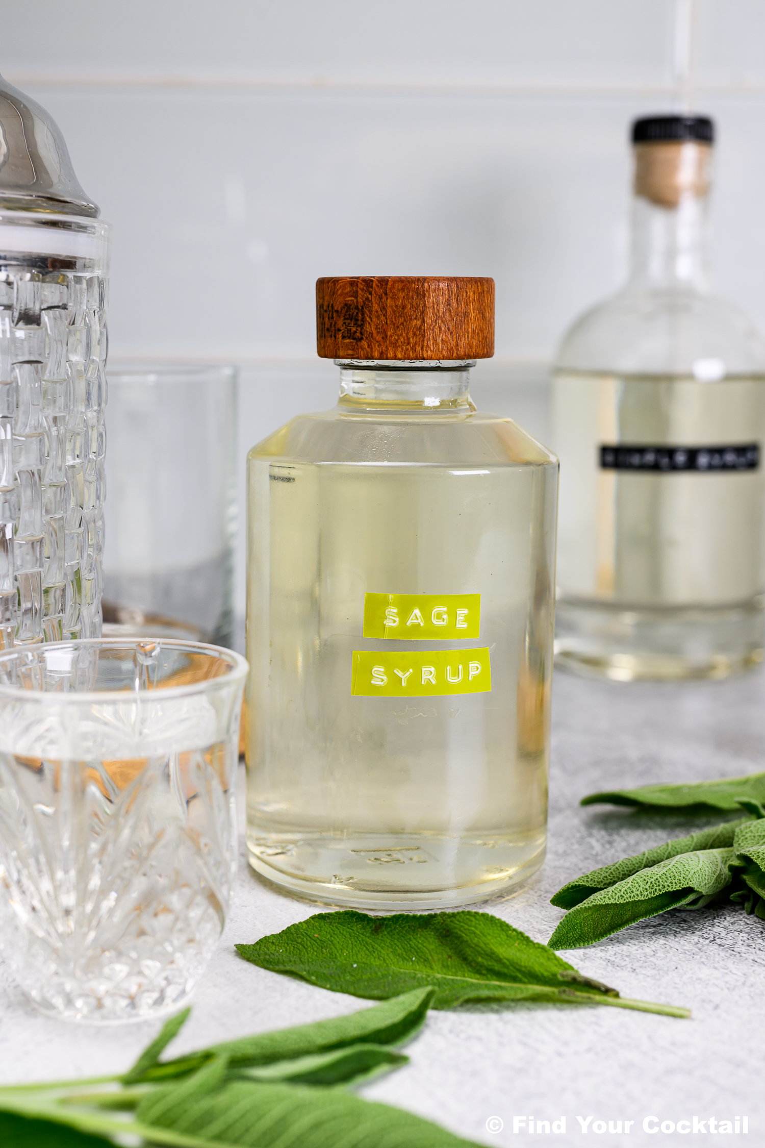 A bottle labeled Sage Simple Syrup sits on a light surface next to sage leaves, a glass, a cocktail shaker, and a wooden board.