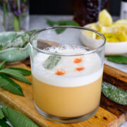 A whiskey sour cocktail in a glass with foam on top, garnished with a green leaf and drops of bitters, sits on a wooden board with lemon wedges and herbs nearby.