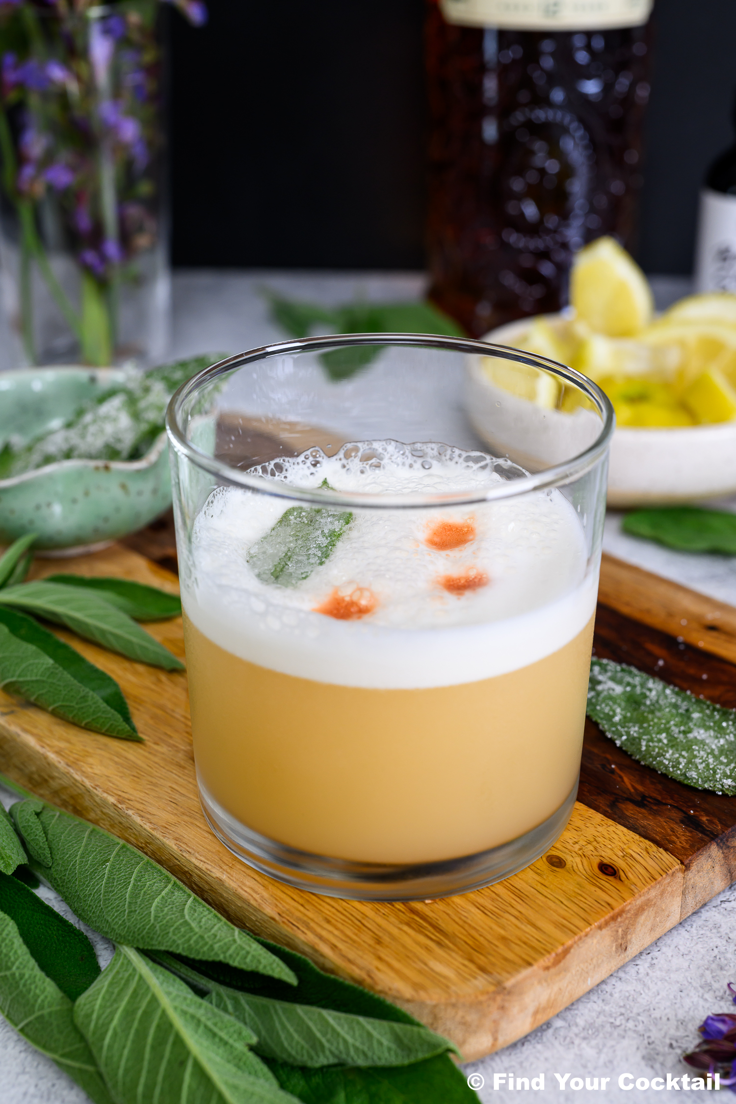A whiskey sour cocktail in a glass with foam on top, garnished with a green leaf and drops of bitters, sits on a wooden board with lemon wedges and herbs nearby.