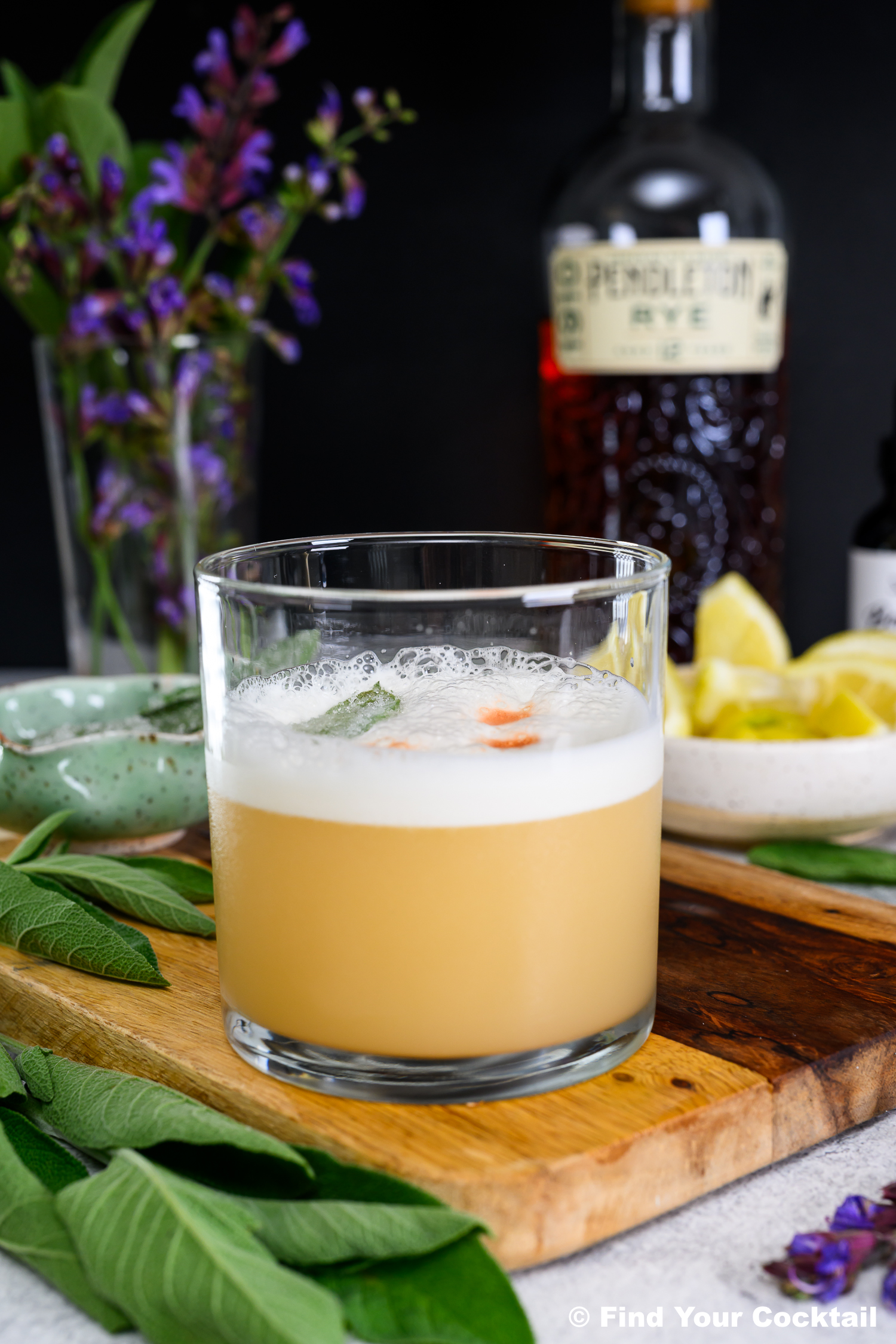 A whiskey sour cocktail in a glass garnished with bitters, set on a wooden board with fresh herbs, lemon wedges, and a bottle of Pendleton rye whiskey in the background.