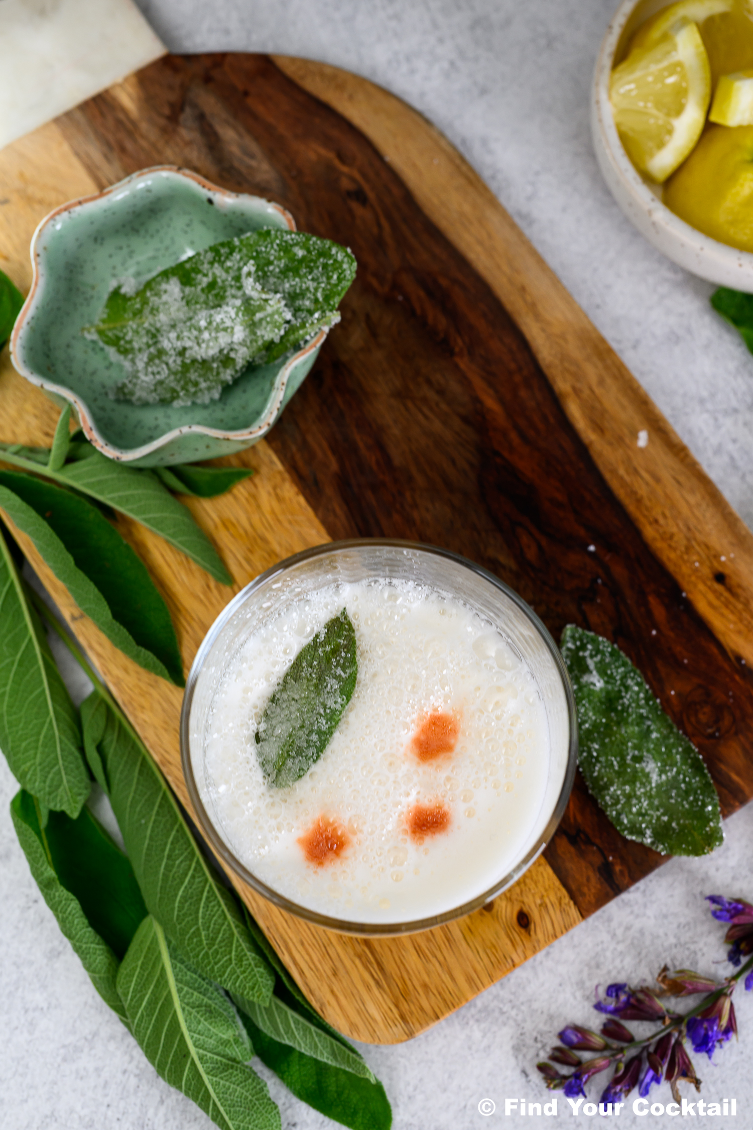 A cocktail with a frothy top, garnished with a sugared sage leaf and three drops of bitters, is placed on a wooden board next to fresh and sugared sage leaves and a bowl of lemon wedges.
