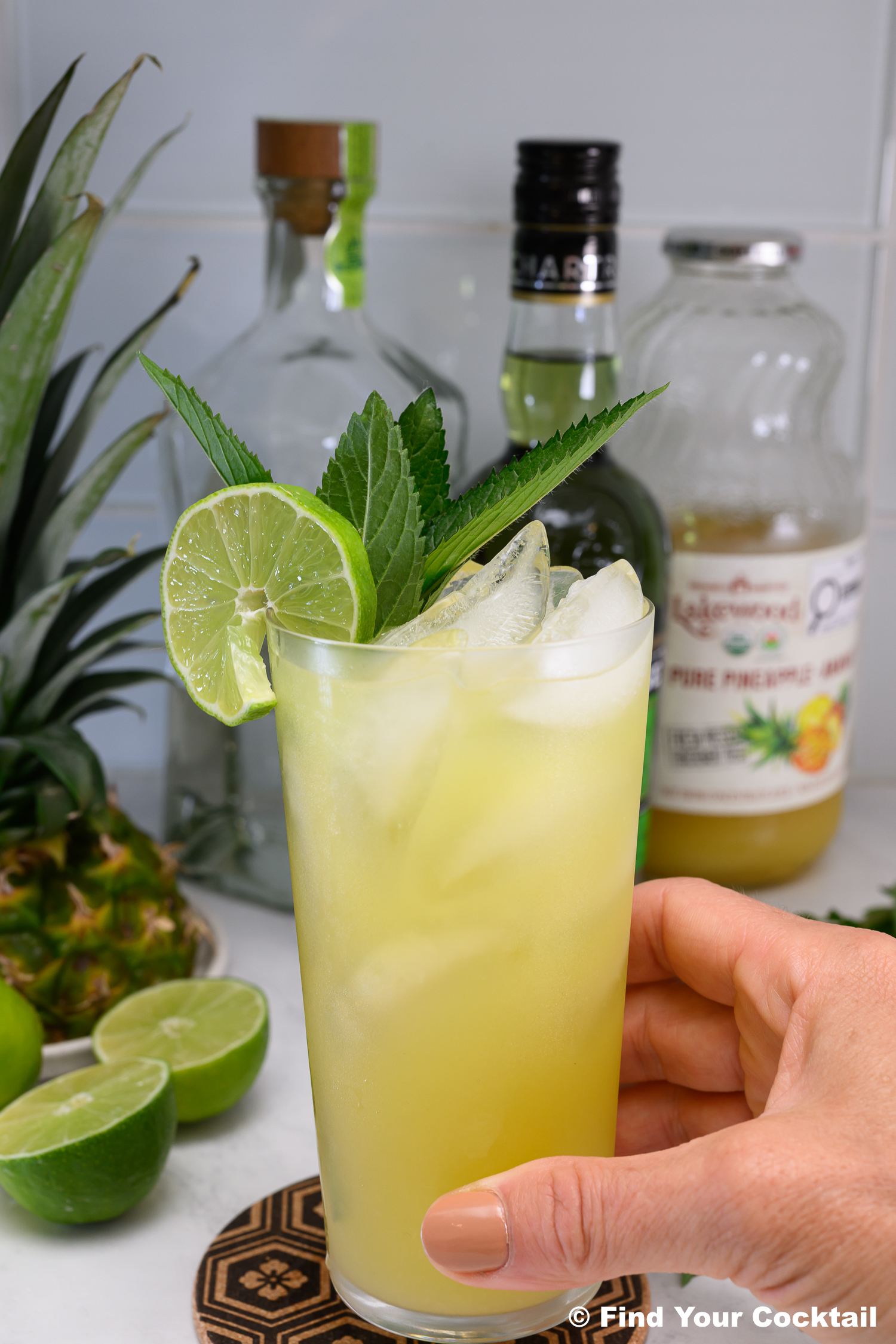 A hand holds a glass of yellow cocktail with ice, lime slices, and mint garnish. Bottles of liquor, pineapple, and pineapple juice are visible in the background.
