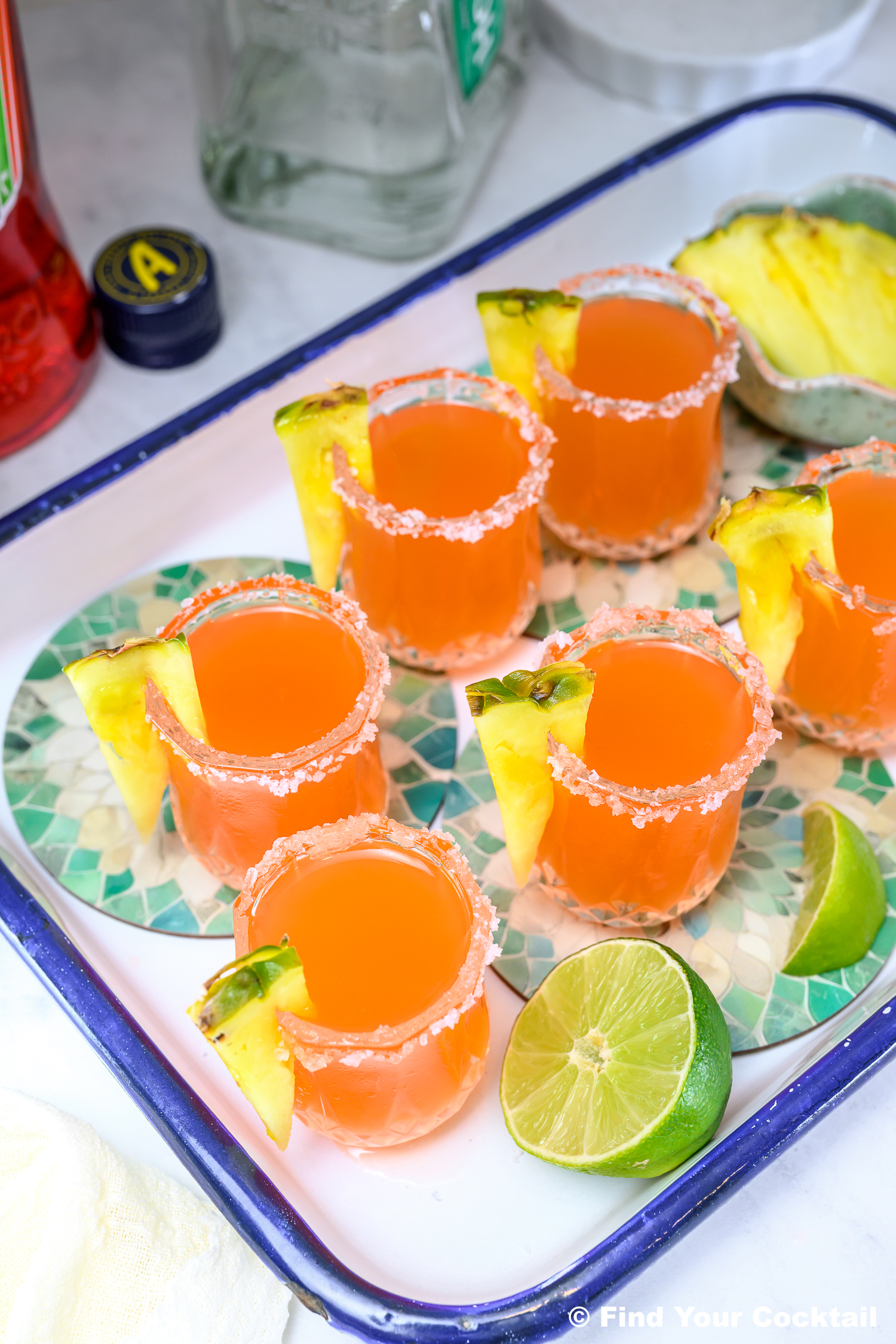 Tray with six orange cocktails in salt-rimmed glasses, each garnished with a pineapple slice; lime halves and a bottle are nearby.