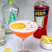 A cocktail with a dried lemon slice garnish in a coupe glass sits on a coaster, surrounded by bottles of Aperol and gin, lemon wedges, and a small bowl of lemon slices.
