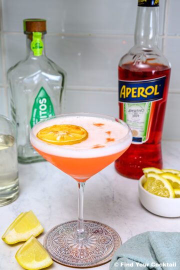 A cocktail with a dried lemon slice garnish in a coupe glass sits on a coaster, surrounded by bottles of Aperol and gin, lemon wedges, and a small bowl of lemon slices.