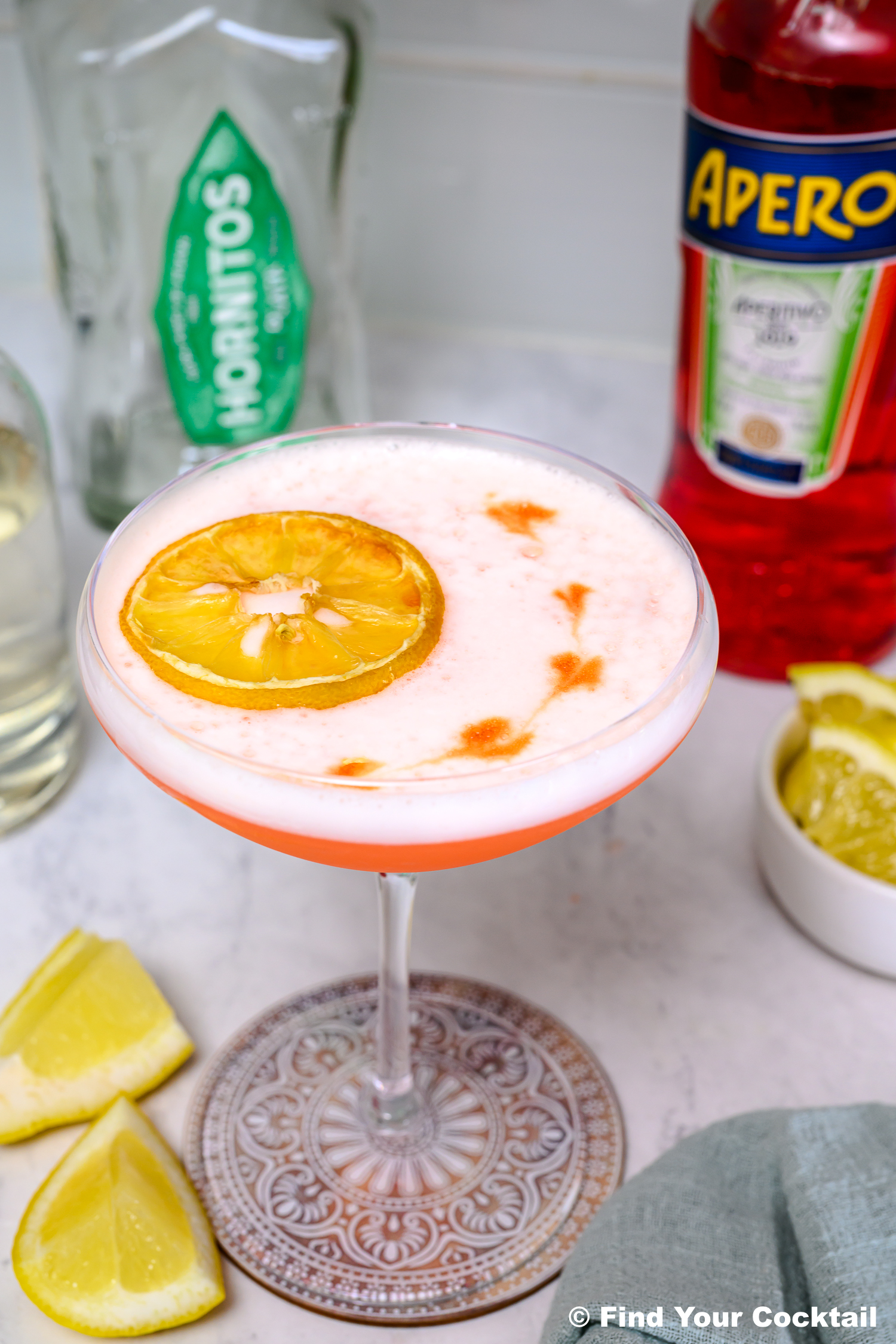 A cocktail in a coupe glass garnished with a dried lemon slice, with bottles of Monin and Aperol, lemon wedges, and a small bowl of limes nearby on a countertop.