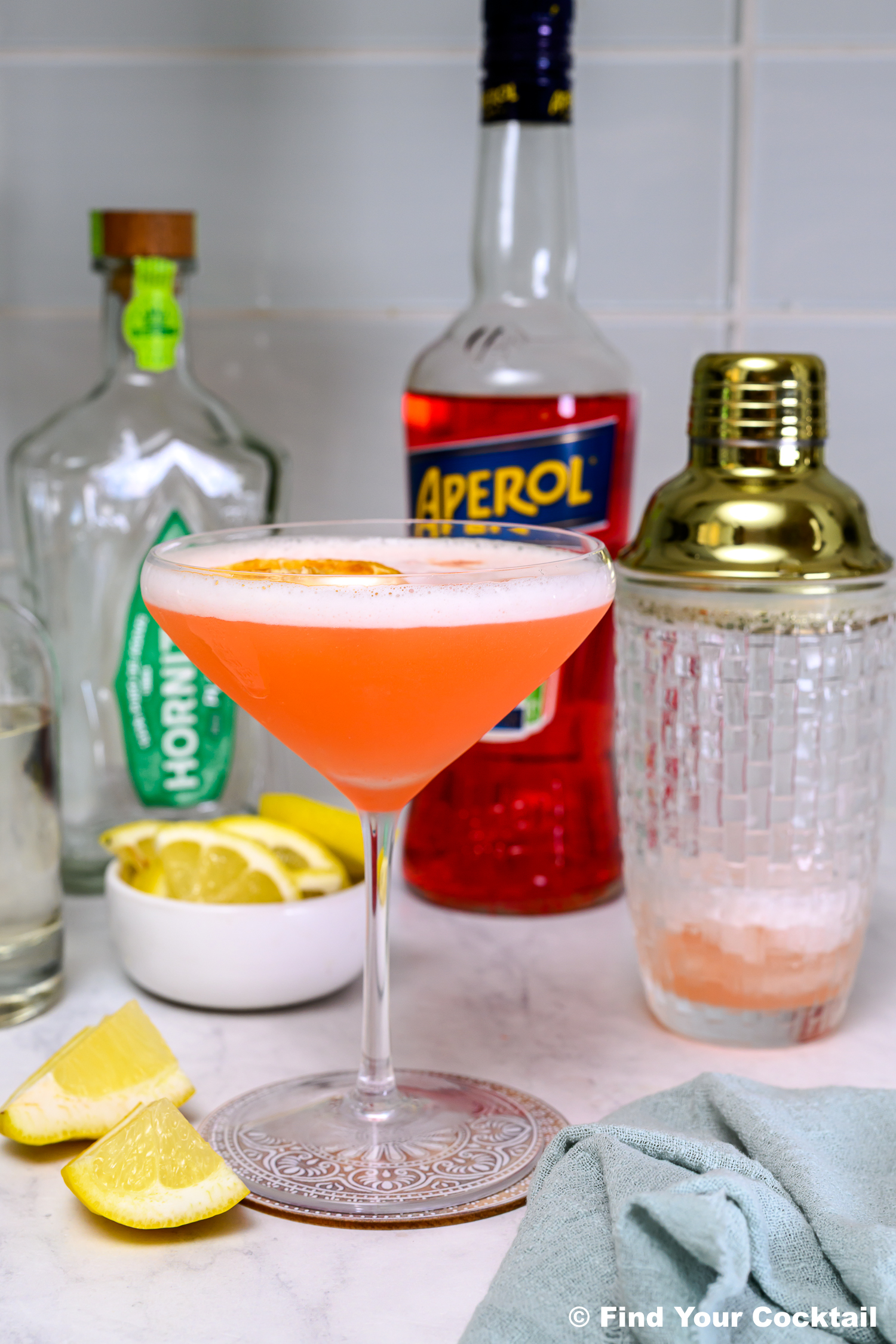 A pink cocktail in a coupe glass garnished with a lemon slice, surrounded by bottles, a cocktail shaker, lemon wedges, and a bowl of lemon slices on a kitchen counter.