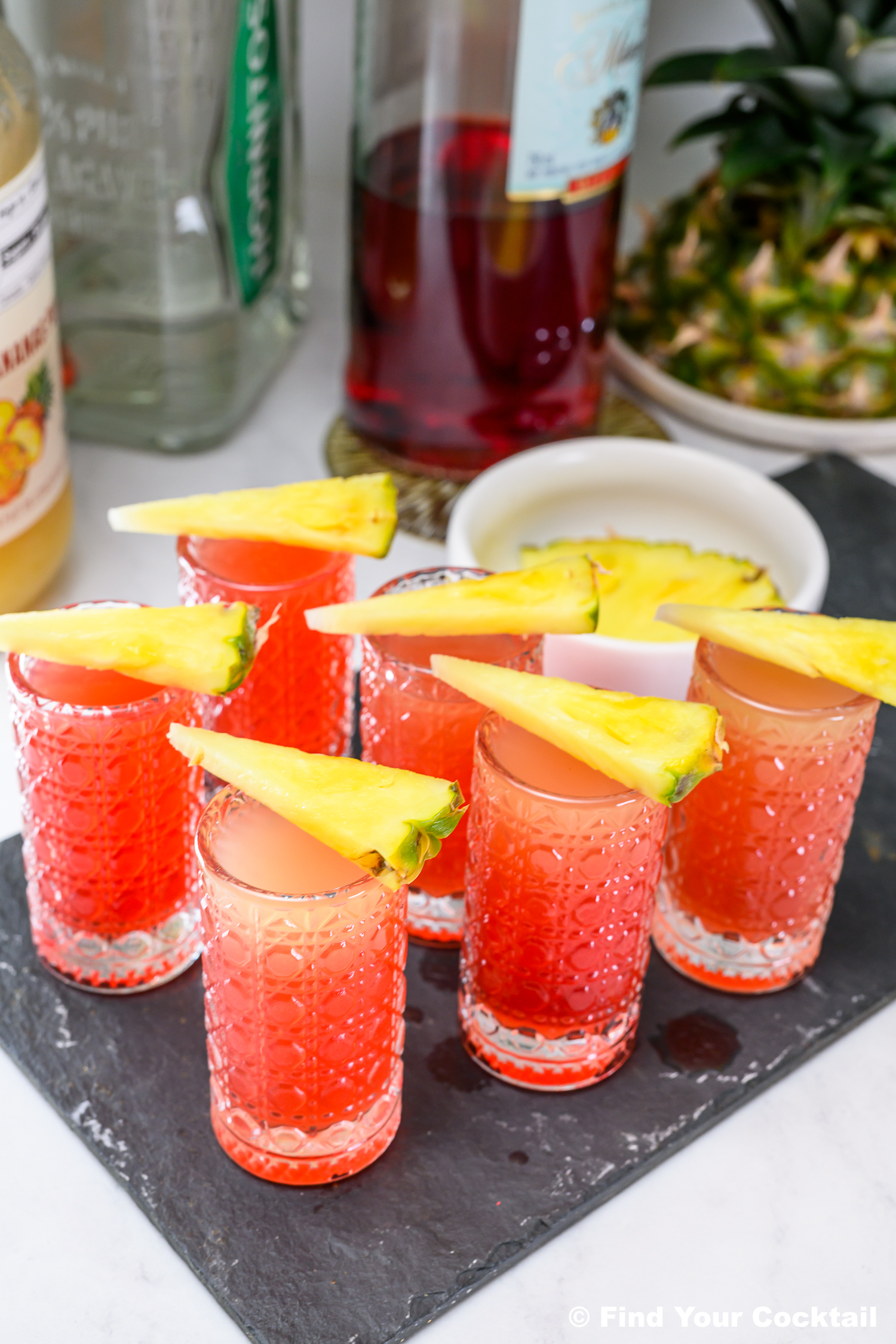 Six red cocktails in textured glasses, each garnished with a pineapple wedge, are arranged on a black tray with bottles and a fresh pineapple in the background.